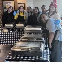Eight smiling women in aprons and blue gloves stand behind buffet food warmers.