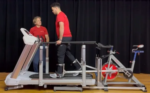 Person walking unassisted on treadmill with other person smiling encouragingly to their right.