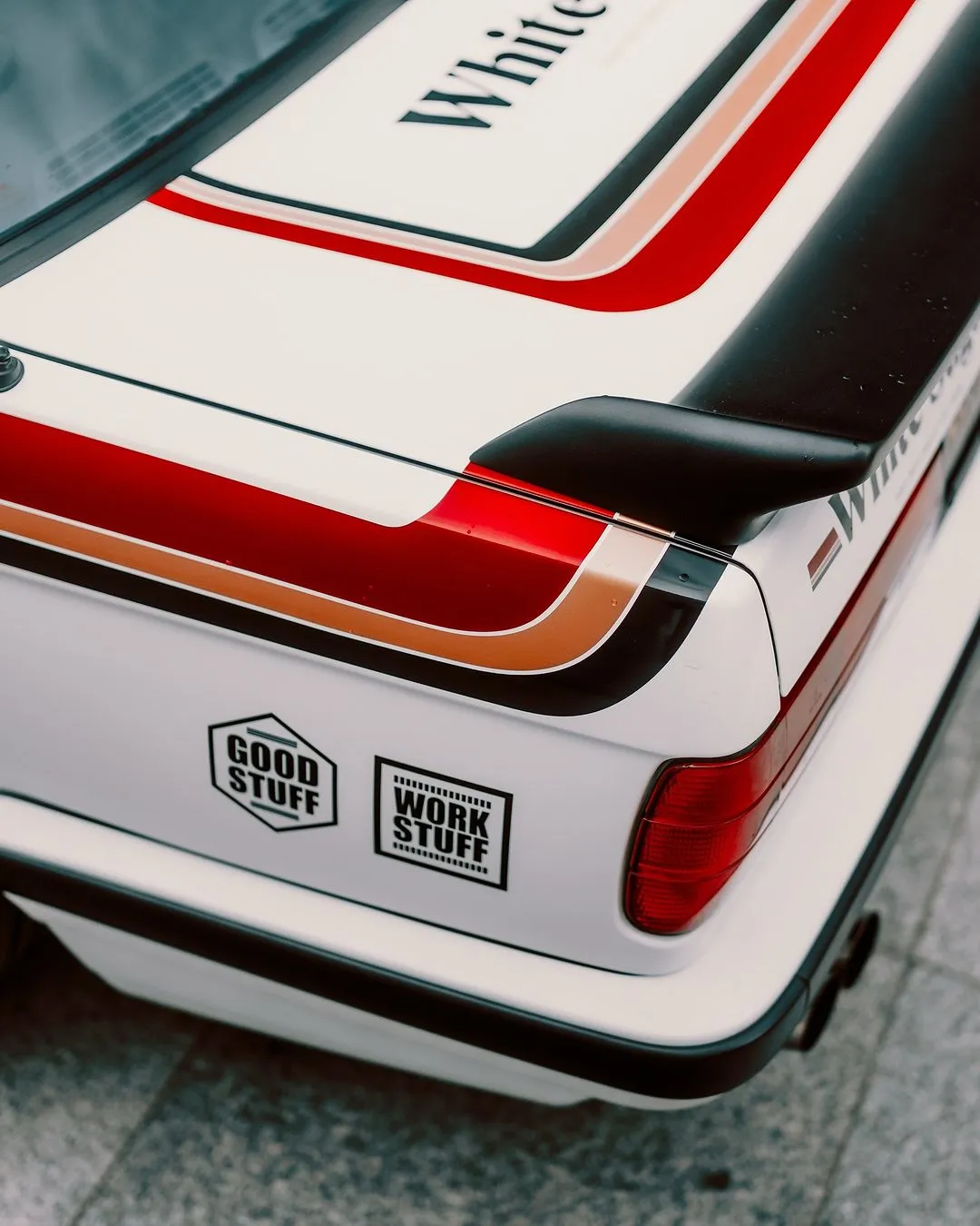 Close-up of the rear corner of a white car with red and brown stripes, a black spoiler, and two stickers that read GOOD STUFF and WORK STUFF. The car is parked on a gray tiled surface.