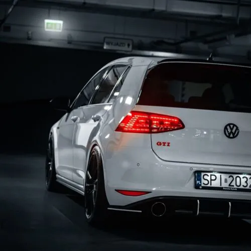 A white Volkswagen Golf GTI with its taillights illuminated is parked in a dimly lit indoor garage, viewed from the rear left side. Part of the license plate is visible, and the car has black wheels—perfect for any Good Stuff team brand ambassador.