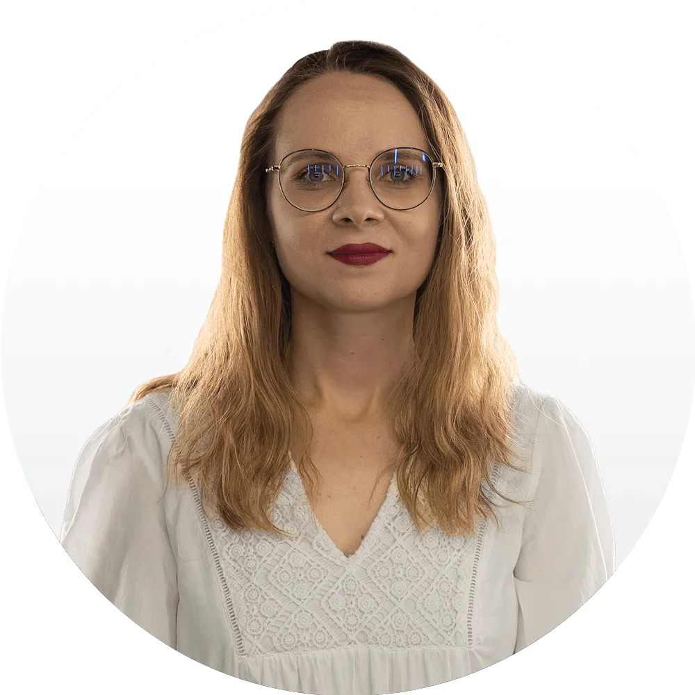 A woman with long, wavy light brown hair, wearing round glasses and a white blouse with lace details, stands in front of a plain white background, looking directly at the camera.