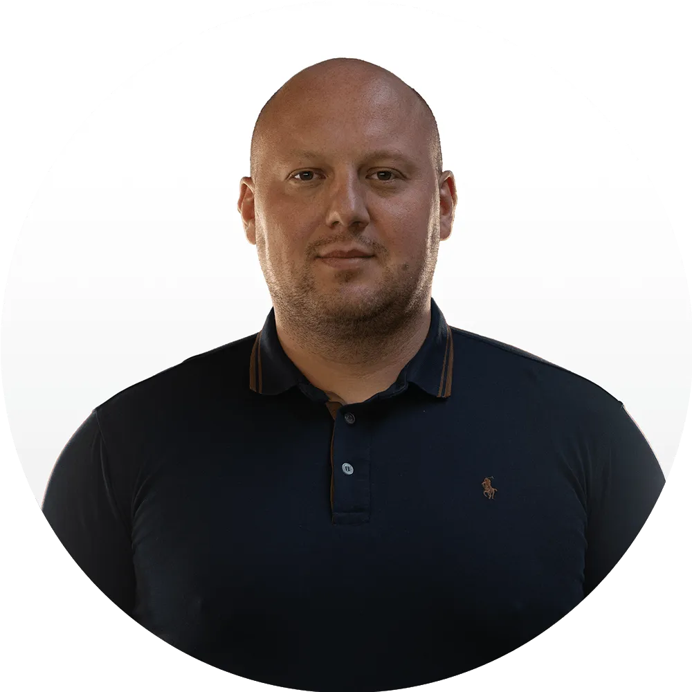 A man with a shaved head wearing a dark navy polo shirt stands against a plain white background, representing the Good Stuff team as he faces the camera with a neutral expression.