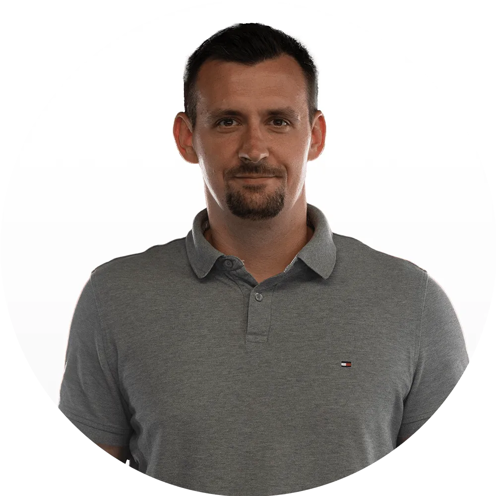 A man with short dark hair and a goatee wearing a gray polo shirt stands against a plain white background, looking directly at the camera with a neutral expression.