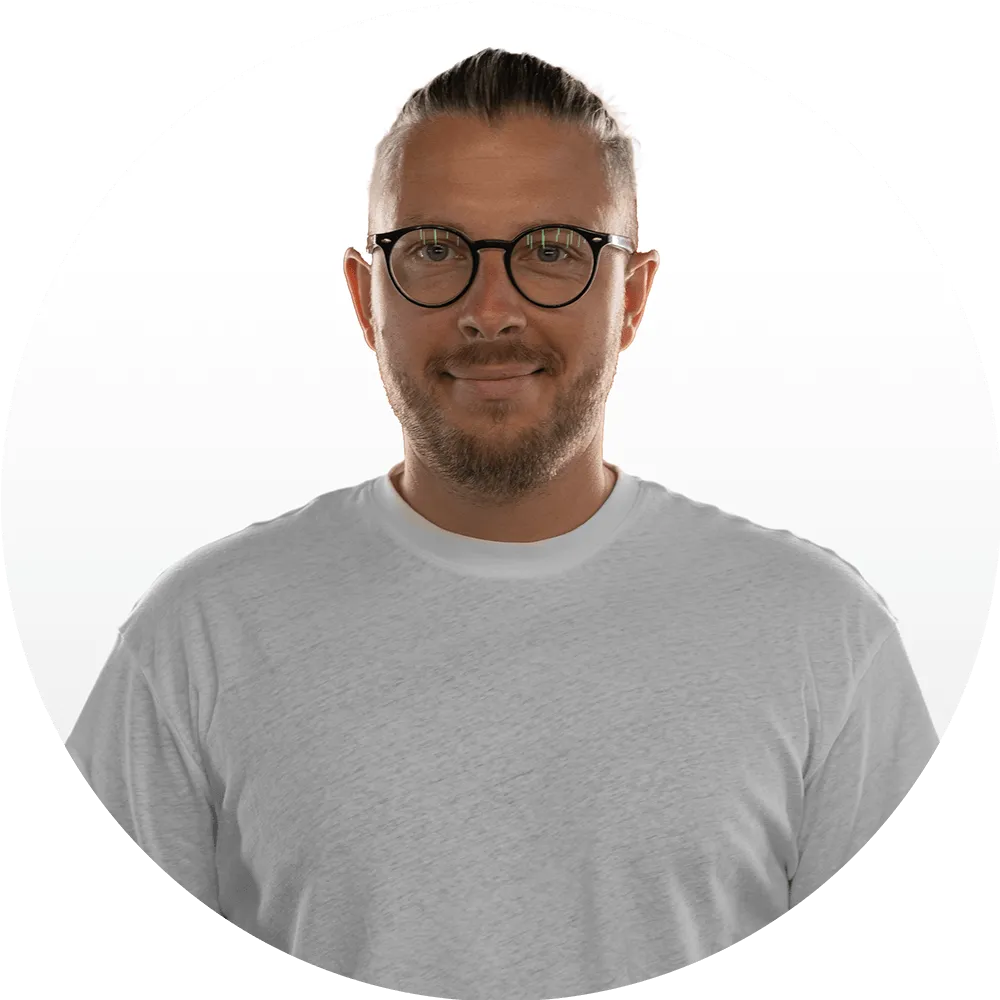 A man with short, slicked-back hair, glasses, and a trimmed beard is wearing a plain white t-shirt and smiling gently at the camera. Meet Us—the Good Stuff team brings warmth and authenticity to our About Us page. The background is white.