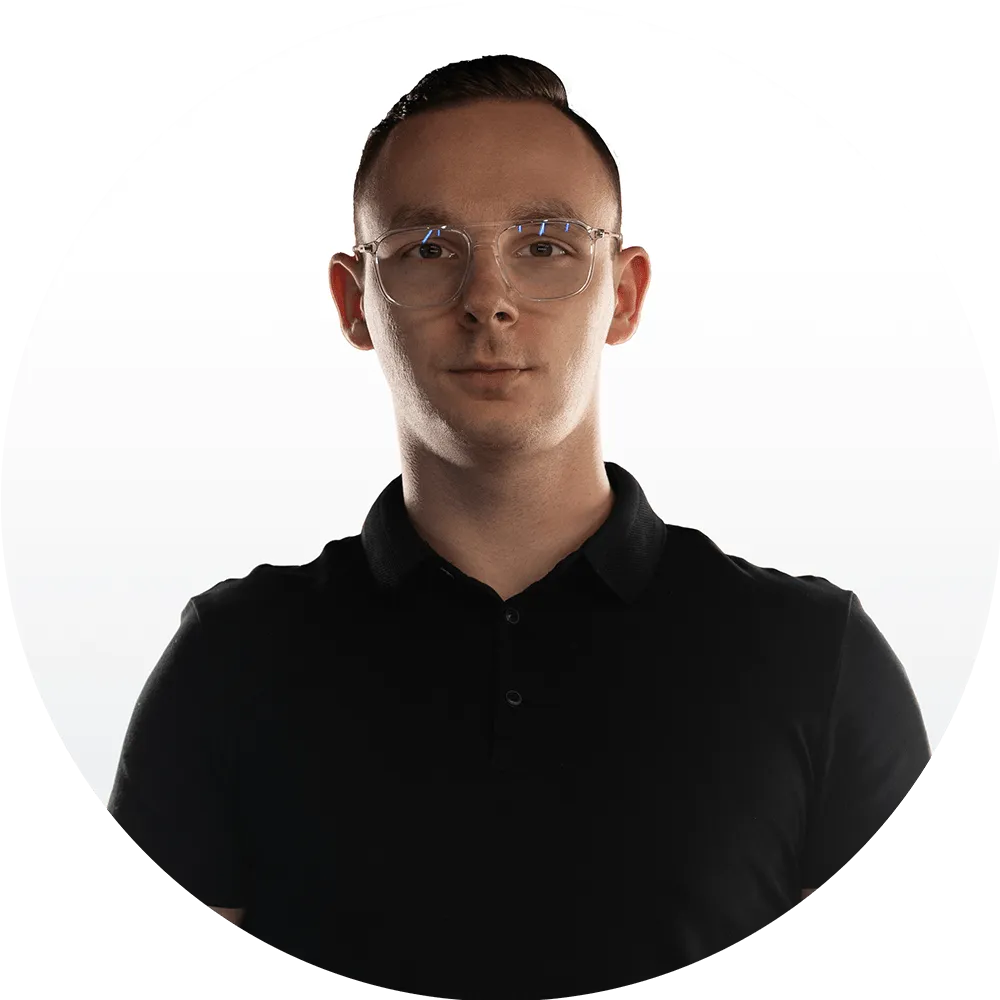A young man with short brown hair, wearing clear eyeglasses and a black collared shirt, stands against a plain white background, looking directly at the camera.