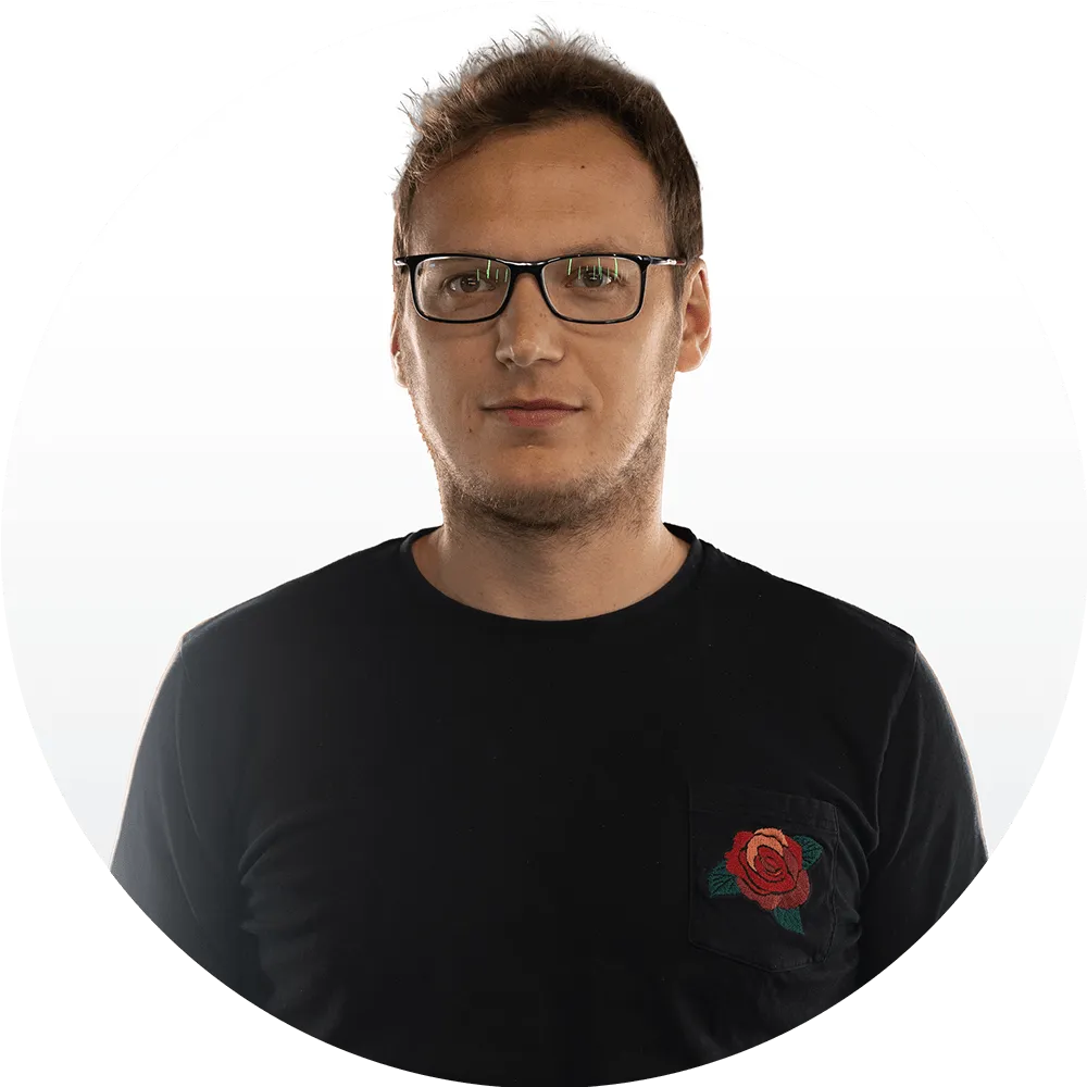 A man with short brown hair and glasses wearing a black shirt with a red rose design on the pocket, poses against a plain white background.