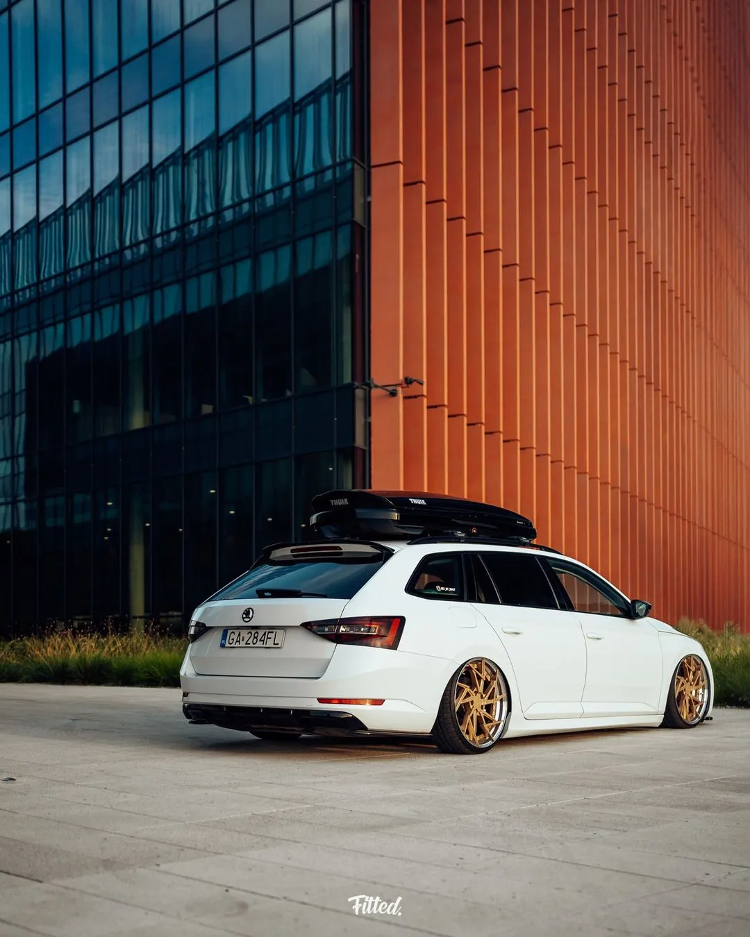 A white station wagon with gold rims and a roof box, representing the Good Stuff team, is parked in front of a modern glass and orange building, captured in bright daylight.