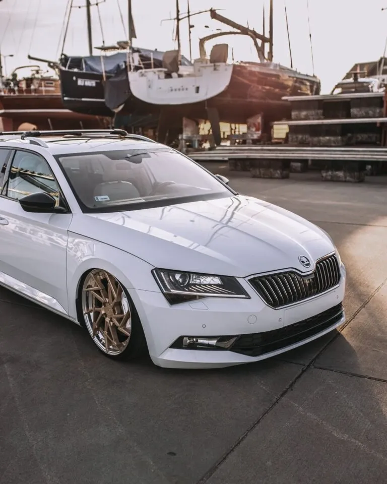 A white Skoda sedan with gold rims is parked on a concrete surface near docked boats and yachts at a marina, sunlight illuminating the scene—perfect for our detailing ambassador. Join us and become part of Project Ambassador today!.