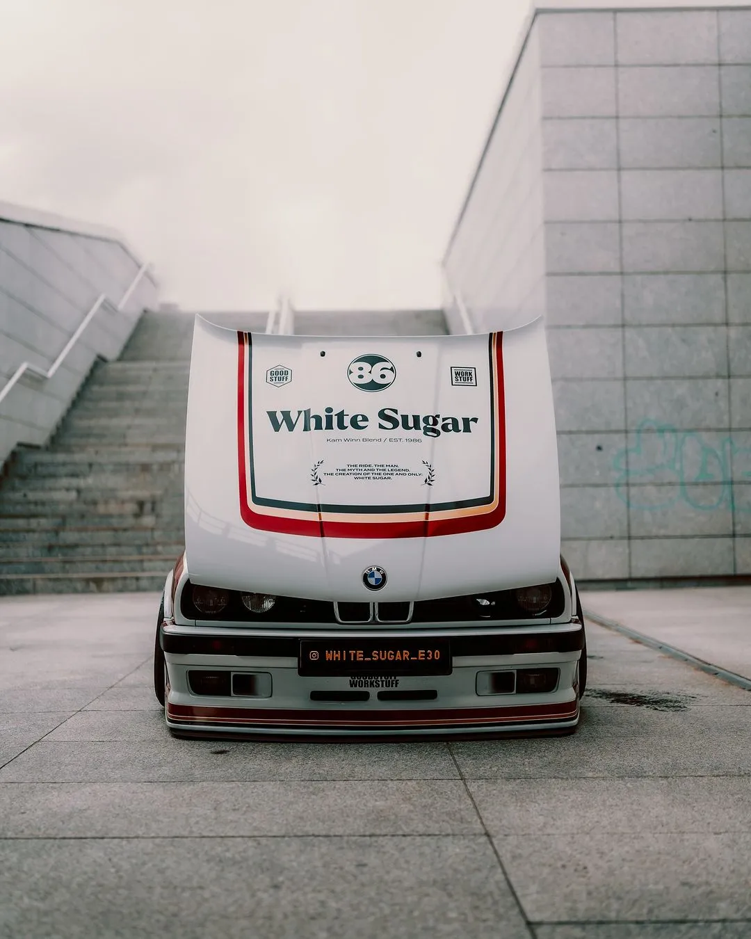 A white BMW car with its hood open, displaying the words White Sugar and the number 36, is parked on a concrete surface near gray stairs and a modern building.