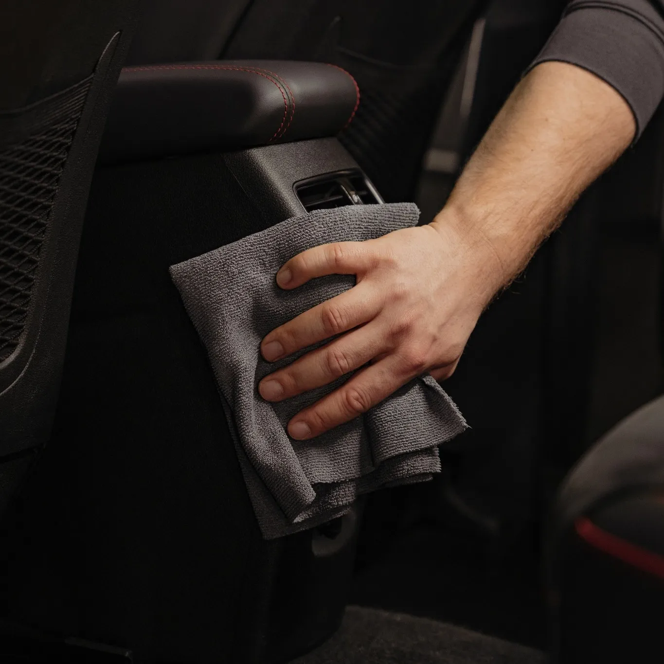 A person focuses on interior detailing by wiping the back of a car seat with a gray microfiber cloth, cleaning the plastic surface near a pocket for that Good Stuff finish.
