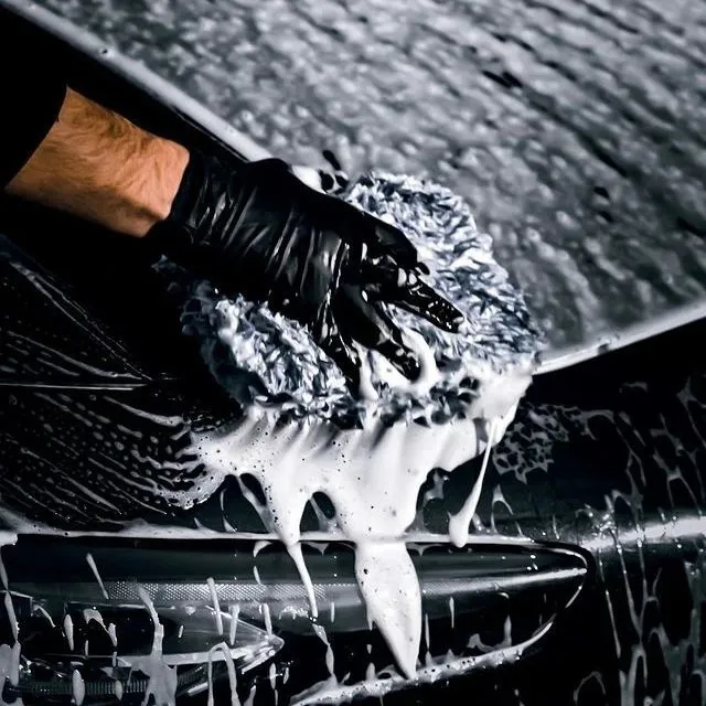 A person wearing a black glove is washing a dark car with a soapy sponge from a car cleaning set, creating white foam that drips down the car’s surface.