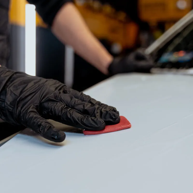 A person wearing black gloves uses a red clay bar to smooth or clean a white surface, with one arm resting on the surface and a blurred background, suggesting a car washing or ceramic coating process.