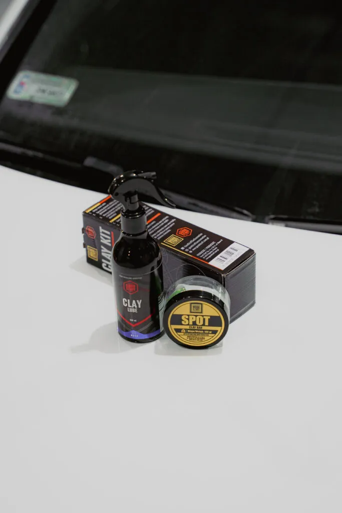 Sobre el capó de un coche blanco, en cajas negras, se exhiben productos de limpieza para coches, entre ellos una botella con la etiqueta Clay Lube y un recipiente redondo con la etiqueta Spot, resaltando la importancia de la descontaminación de la pintura de coche.