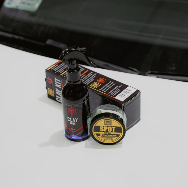 A bottle of clay lube spray and a tin labeled Spot sit on a white car hood, perfect for car washing or prepping for ceramic coating, with product boxes and a windshield visible in the background.