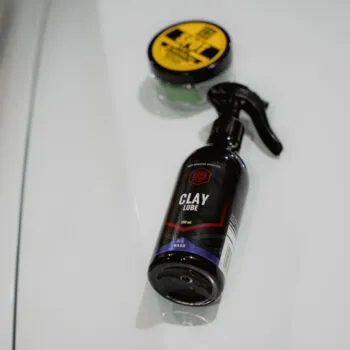 A black spray bottle labeled “Clay Lube” and a round container with a yellow lid—perfect for clay bar paint decontamination—rest on a glossy white surface, likely the hood of a car.