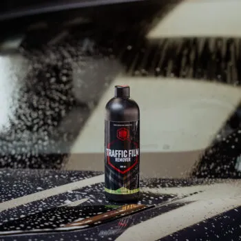 A bottle of Traffic Film Remover, ideal for paint decontamination, is placed on the hood of a car covered in soap suds and water droplets, with the windshield visible in the background.
