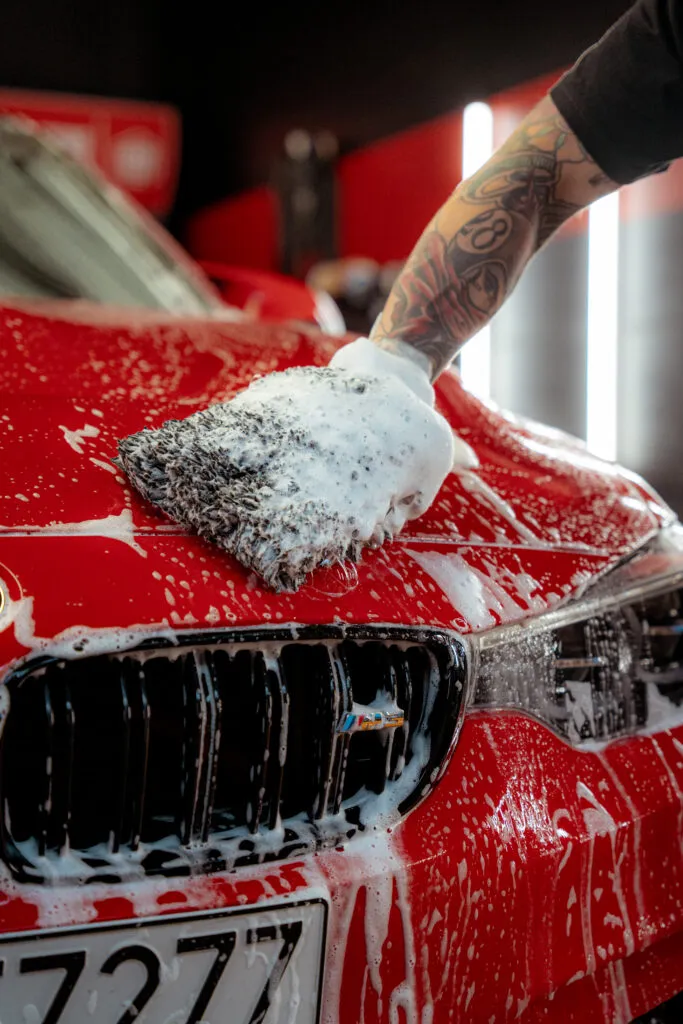 Un brazo tatuado utiliza una manopla jabonosa para limpiar el capó de un coche rojo brillante, centrándose en el prelavado para proteger la pintura, con espuma que cubre la parrilla delantera del coche y la zona de los faros.