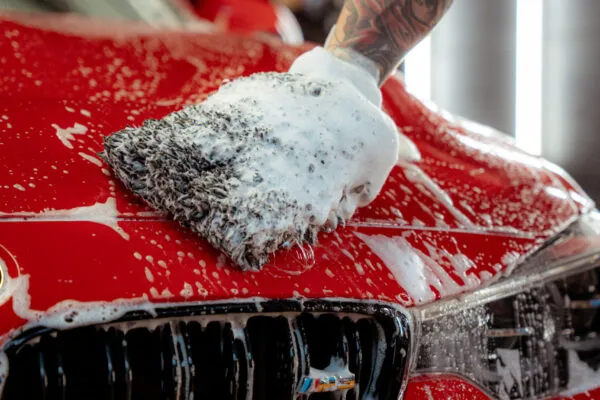 Ein tätowierter Arm benutzt einen seifigen Waschhandschuh, um die Motorhaube eines roten Autos zu reinigen, wobei er sich auf den Bereich des Kühlergrills konzentriert. Dicke weiße Seifenlauge tropft als Teil der Vorwäsche herunter und hilft, Lack zu schützen während dieser gründlichen Autodetailbehandlung.