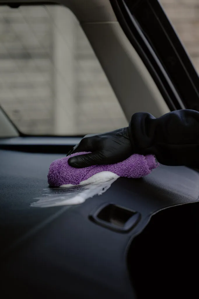 A person wearing a black glove wipes the dashboard of a car with a purple microfiber cloth, giving attention to detail after using upholstery cleaner for a thorough clean car seat and interior finish.
