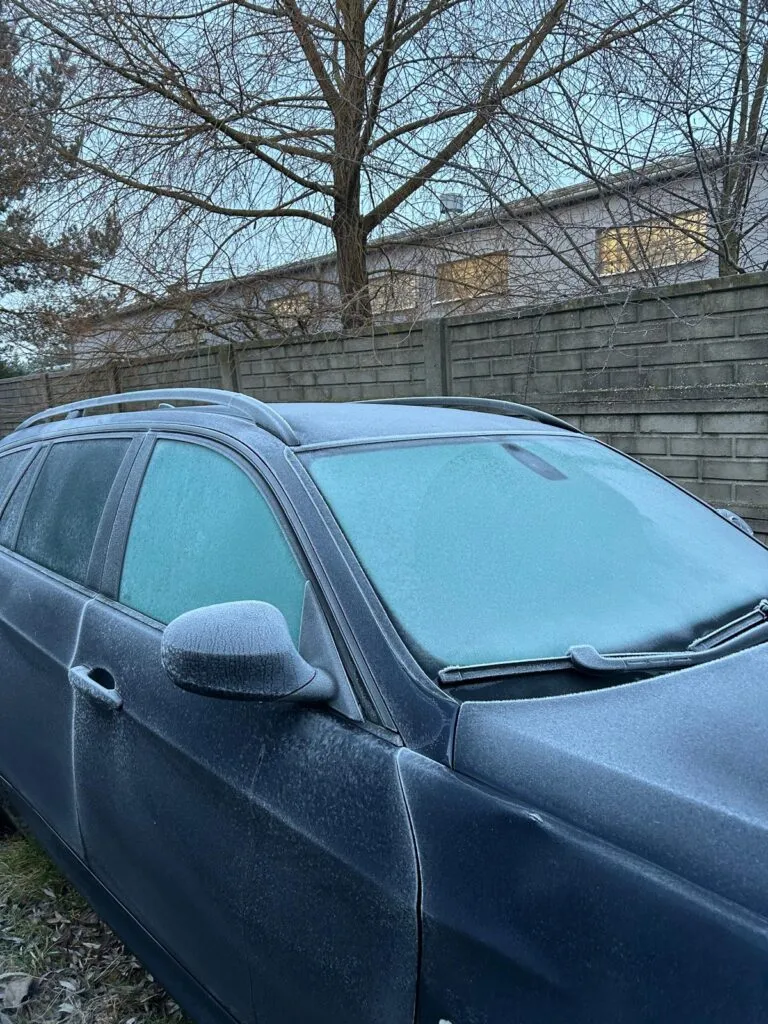 A dark-colored car is parked outdoors, covered in frost on the windows and roof. Leafless trees and a stone wall are in the background, with a building visible behind the wall, hinting at the need to safely remove ice from the car on a cold winter morning.
