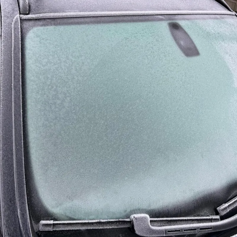 A car windshield covered in a layer of frost, with windshield wipers resting at the bottom and frost also visible on the roof and hood—highlighting the need to safely remove ice from your car. A concrete wall is in the background.