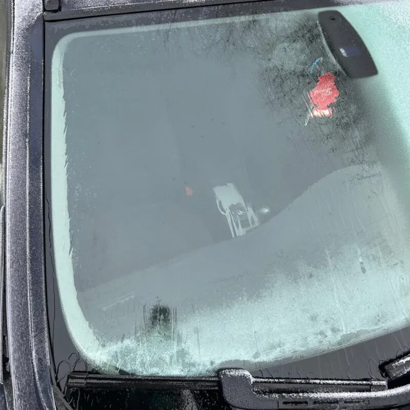 A car windshield partially covered in frost, with the wipers clearing a section of the glass—a classic scene of car ice removal. A red air freshener hangs from the rearview mirror inside the car.