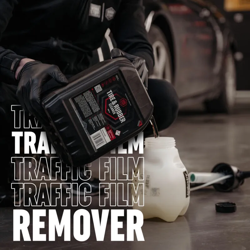 Eine Person mit schwarzen Handschuhen schüttet eine Flüssigkeit aus einem schwarzen Behälter mit der Aufschrift Tire & Rubber Cleaner in einen weißen Plastikkrug. Der Schriftzug TRAFFIC FILM REMOVER ist in fettgedruckter Schrift zu sehen.