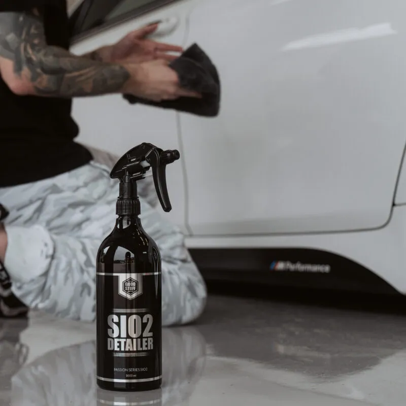 Eine Person kniet neben einem weißen Auto und hält ein schwarzes Tuch in der Hand, um die Tür zu reinigen. Im Vordergrund steht eine Sprühflasche mit der Aufschrift SiO2 Detailer auf einem glänzenden Boden.