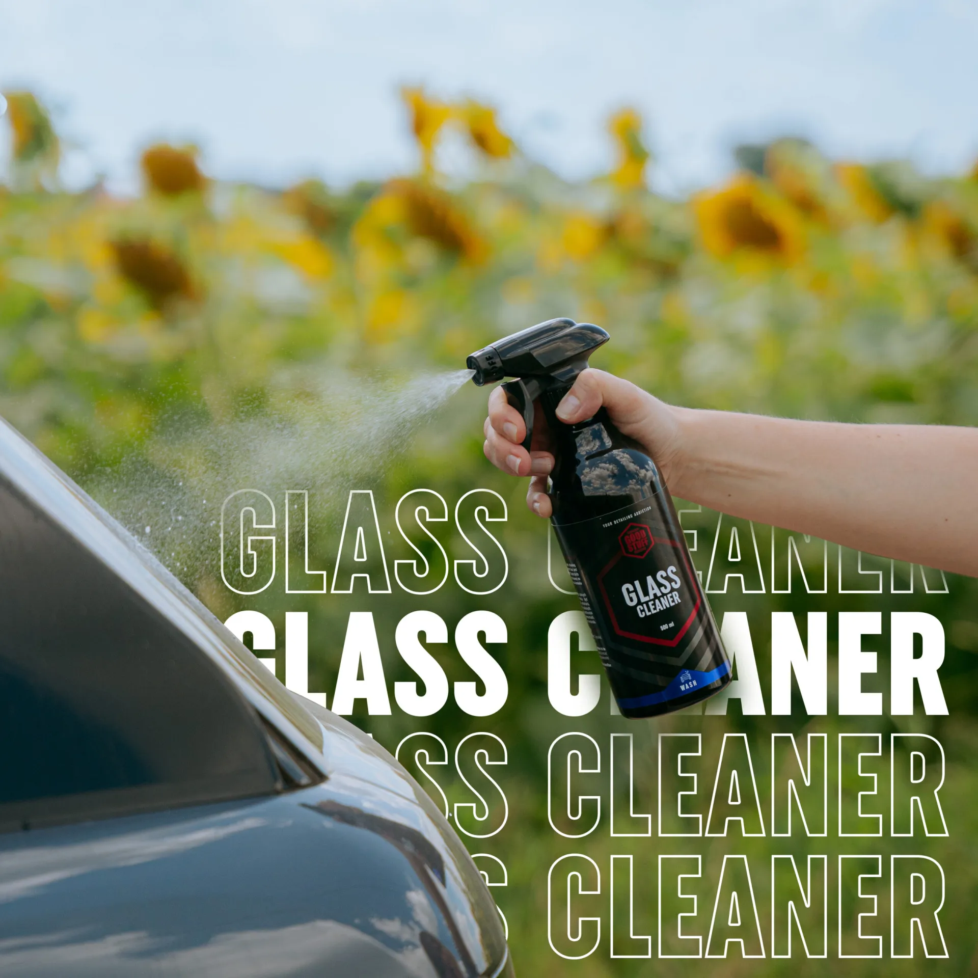 Ręka rozpylająca płyn do mycia szyb na szybę samochodu na zewnątrz, ze słonecznikami i zielenią w tle. Słowa GLASS CLEANER są wielokrotnie nakładane pogrubionym białym tekstem.