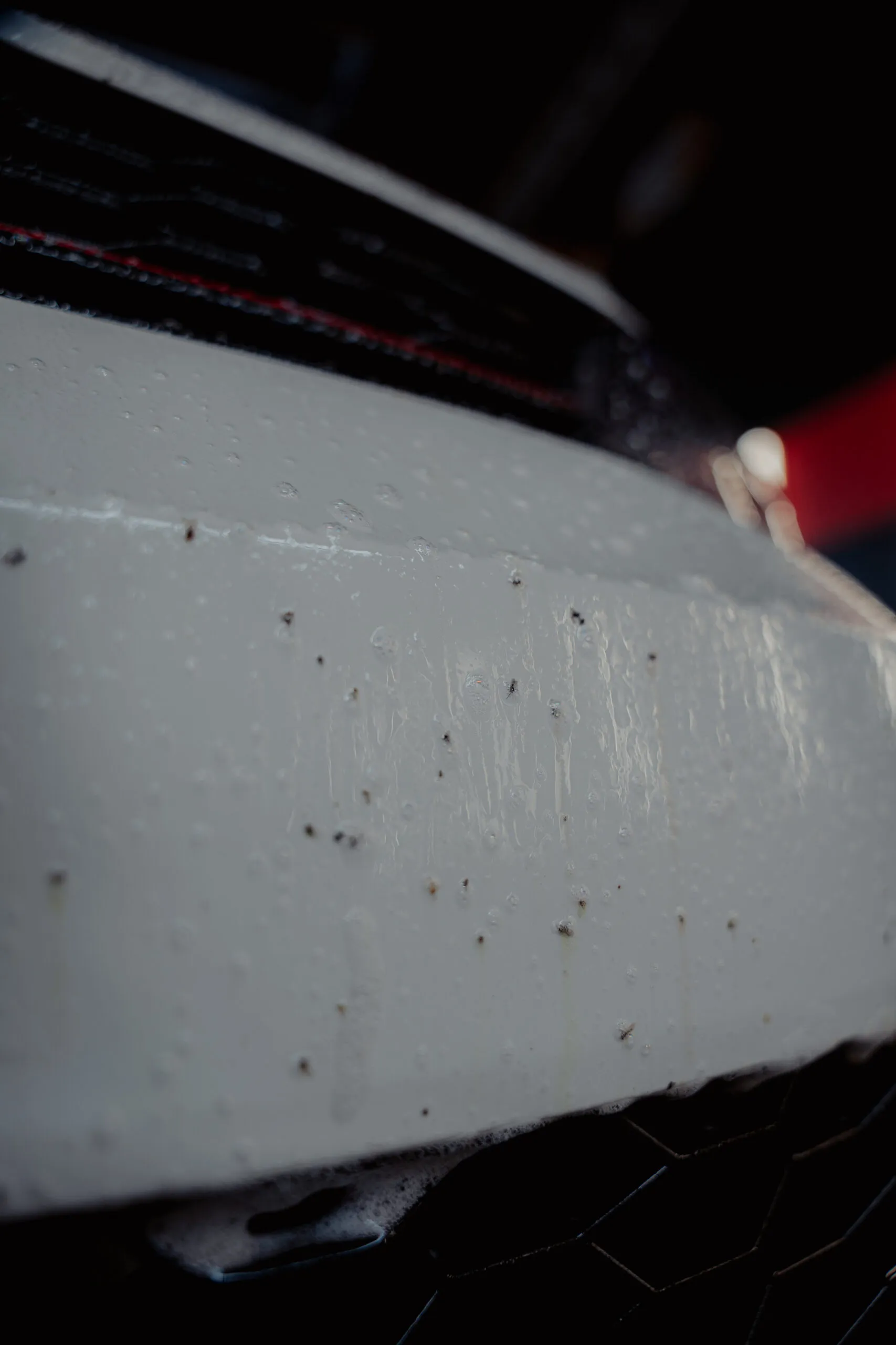 Close-up of a white car's front bumper covered in water droplets and small dead insects after a drive—perfect scene to put Good Stuff Bug Buster car bug remover to the test, with blurred background details.