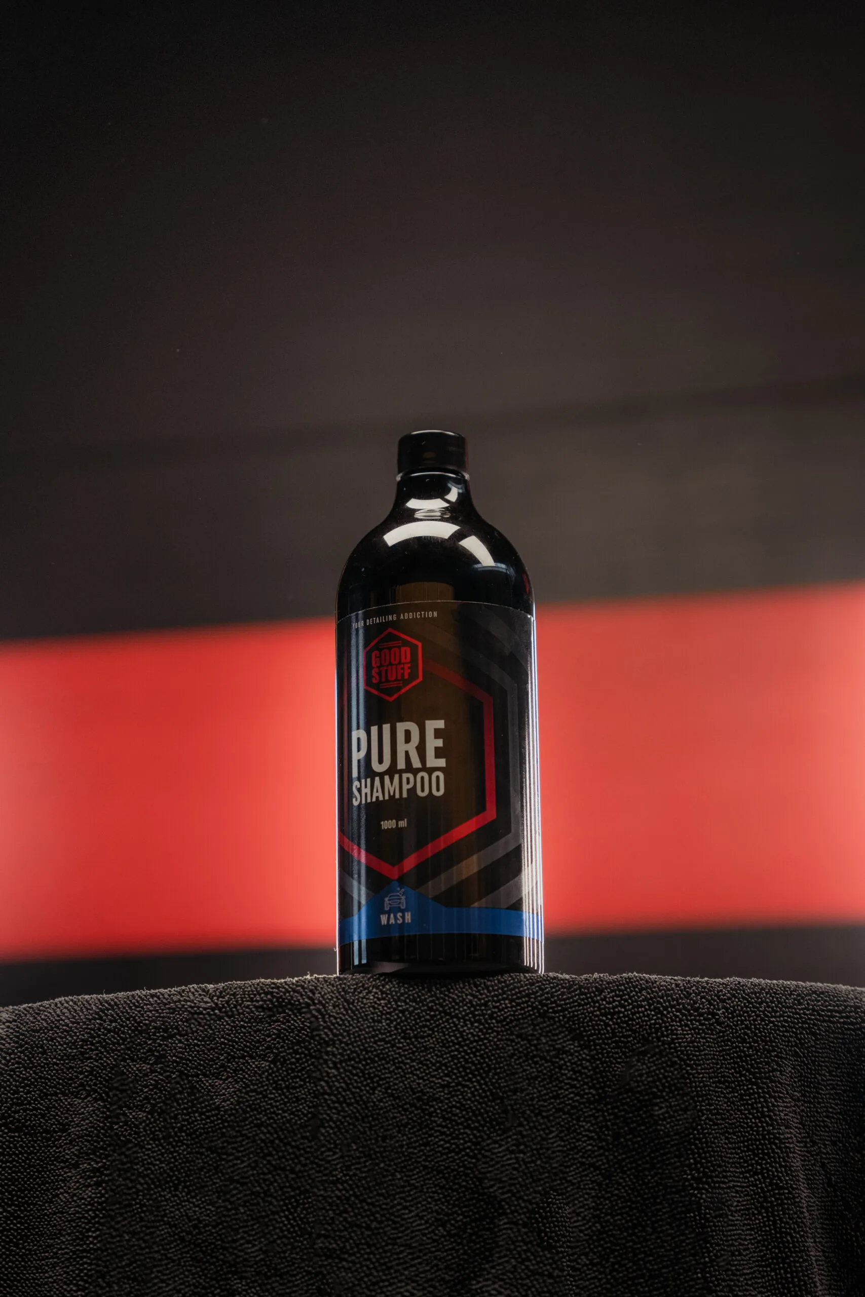 A black bottle of Pure Shampoo by Good Vibes, also known as a premium car shampoo, sits on a dark towel, with a blurred red and black background.