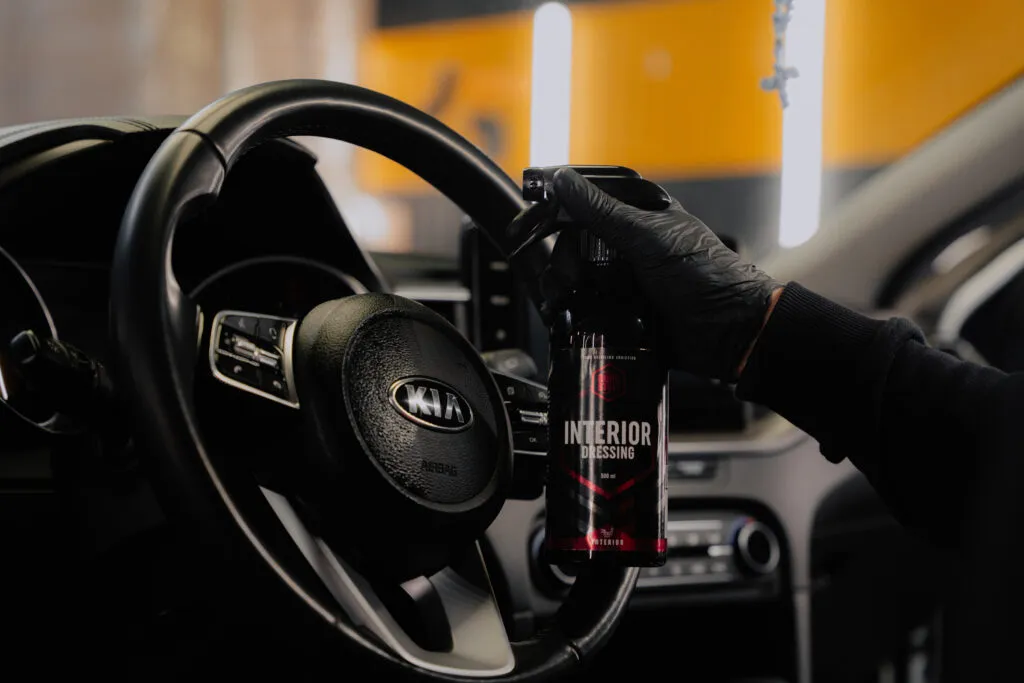 A gloved hand holds a spray bottle labeled Interior Dressing next to a Kia car steering wheel inside a vehicle, suggesting car interior cleaning or detailing.
