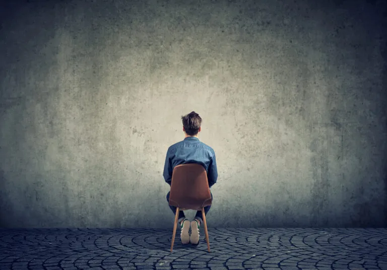 Eine Person sitzt rückwärts auf einem Stuhl, die Last der Depression ist spürbar, während sie auf eine kahle, strukturierte Wand auf einem Kopfsteinpflaster blickt.