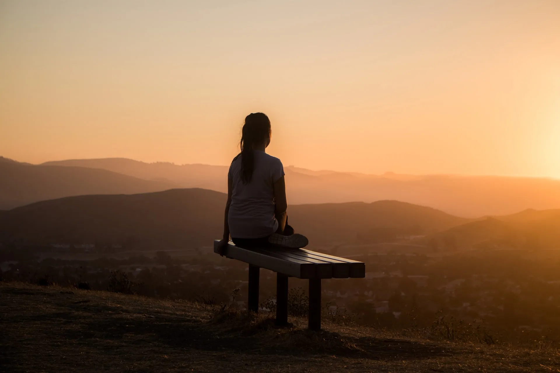 Person, die bei Sonnenuntergang auf einer Bank sitzt und auf eine malerische Landschaft mit Hügeln und einem dunstigen Himmel im Hintergrund blickt.