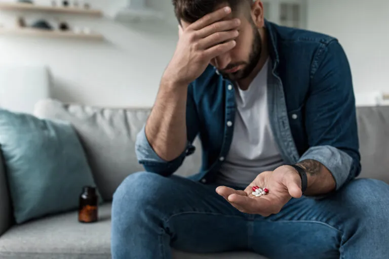 Ein Mann sitzt auf einem Sofa, hält in einer Hand mehrere Antidepressiva-Tabletten und ruht mit der Stirn auf der anderen. Daneben steht eine Tablettenflasche.