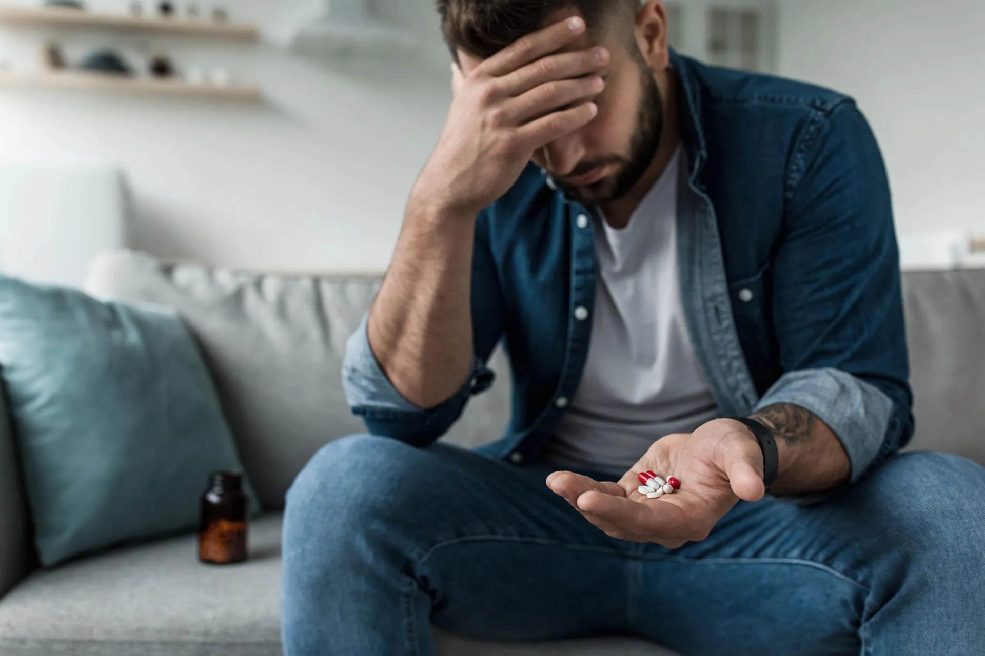 Ein Mann sitzt auf einem Sofa, hält in einer Hand mehrere Antidepressiva-Tabletten und ruht mit der Stirn auf der anderen. Daneben steht eine Tablettenflasche.