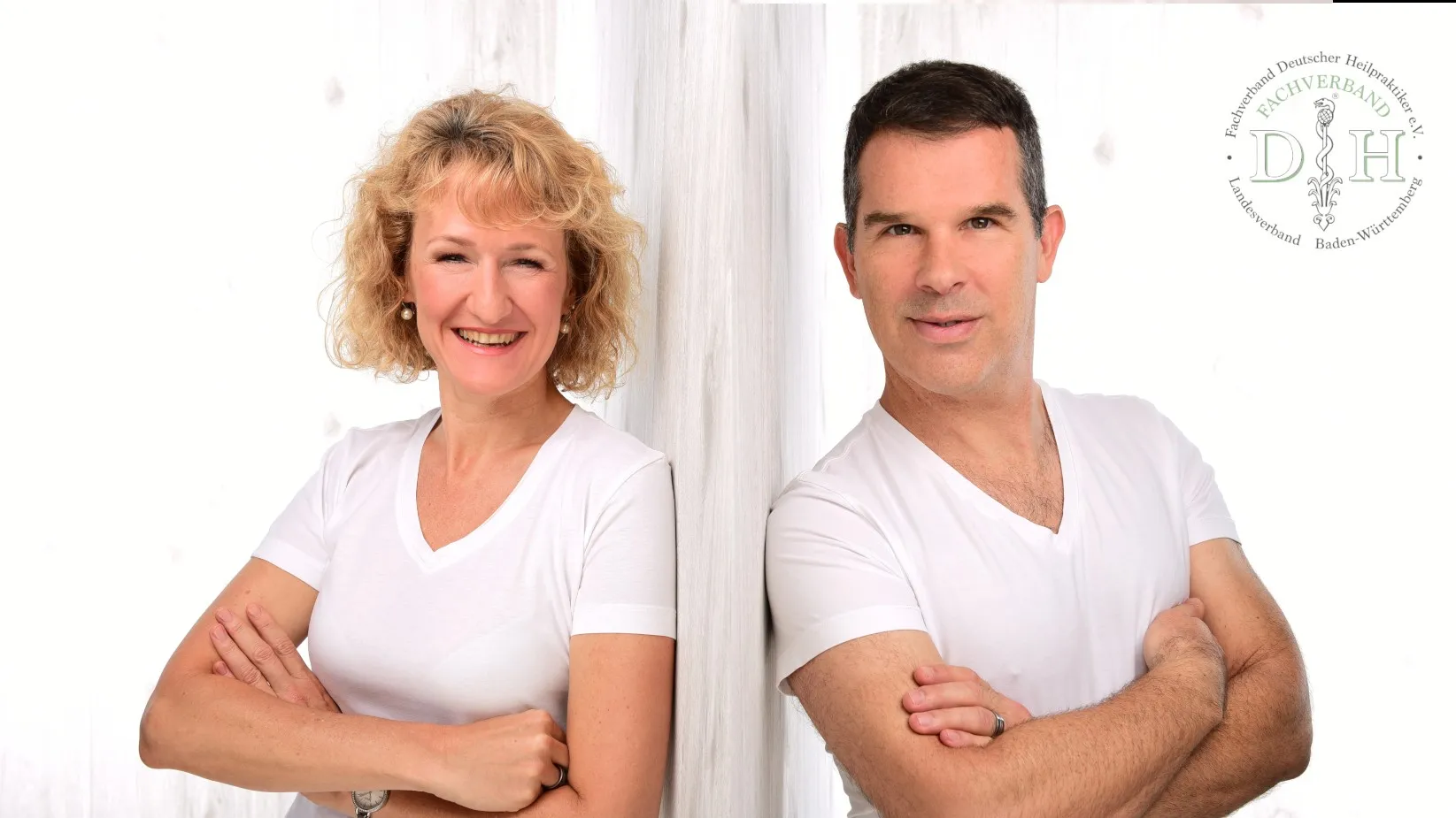 Eine Frau und ein Mann stehen Rücken an Rücken mit verschränkten Armen, beide tragen weiße T-Shirts vor einem weißen Hintergrund mit einem Logo in der oberen rechten Ecke – was ihr gemeinsames Engagement für Homöopathie und alternative Heilmethoden als Heilpraktiker Tübingen widerspiegelt.