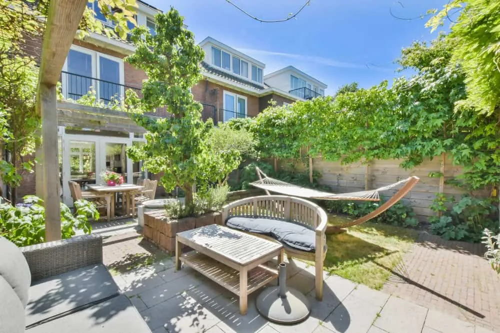Een zonnige achtertuin met weelderig groen, een hangmat, een houten bank met kussens en een salontafel op een stenen patio. Er is een pergola, een eettafel en stoelen bij glazen deuren die leiden naar een stenen huis met grote ramen en balkons erboven.