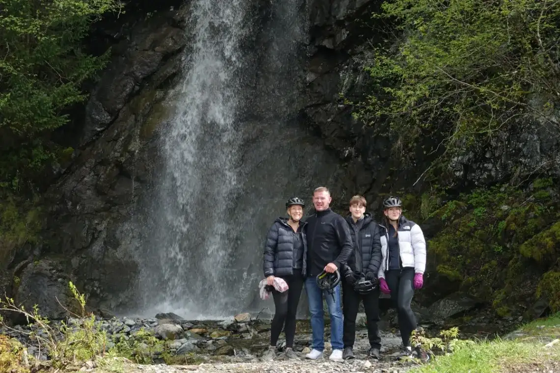 Eco-Friendly Tours in Juneau Alaska: Travel Green with iRide 1 Four iRide Alaska guests with bike helmets smiling in front of a powerful waterfall on an eBike tour.