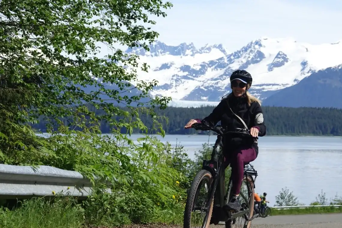 Juneau-rainforest-ebike-woman-smile-glacier-ocean-forest-view-1200x800 Smiling woman on a Juneau eBike tour with snow-capped mountains and Mendenhall Glacier.