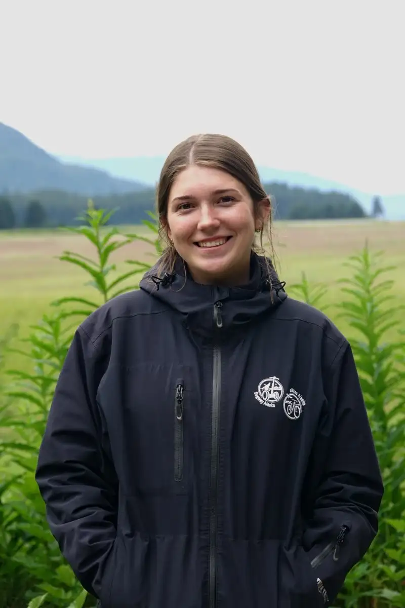 Get to Know Us 10 A smiling woman wearing a black jacket stands outdoors in a scenic green field.