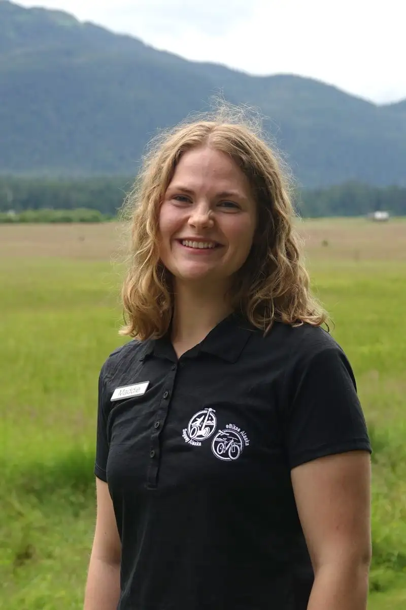 Get to Know Us 3 Smiling blonde woman wearing a black polo shirt with green mountains in the background.