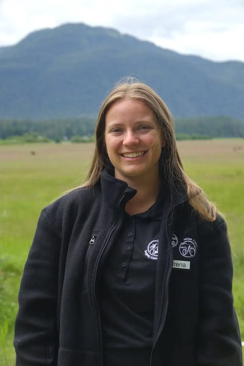 Get to Know Us 6 A smiling woman wearing a black fleece jacket stands outdoors against a mountain backdrop.