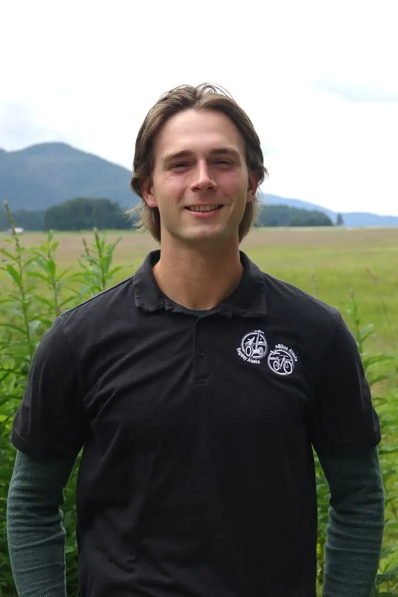 Get to Know Us 5 Smiling young man in a black polo shirt standing outdoors with mountains behind him.