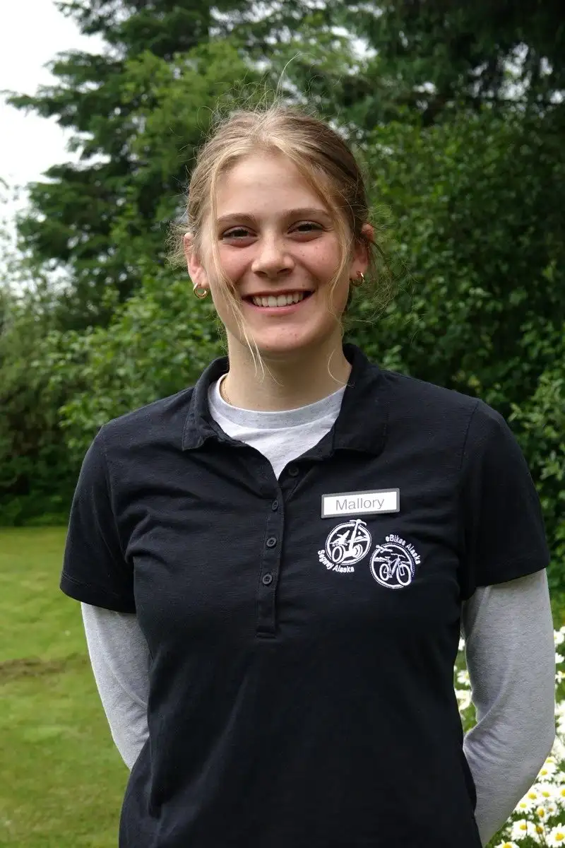 Get to Know Us 11 A smiling young blonde woman named Mallory wearing a black polo shirt outdoors.