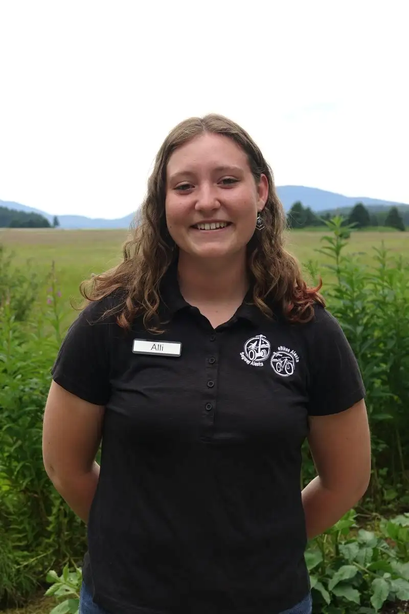 Get to Know Us 8 A smiling woman named Alli wearing a black polo shirt outdoors in a grassy field.