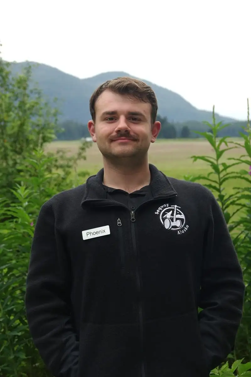 Get to Know Us 7 A smiling man named Phoenix wearing a black fleece jacket outdoors with mountains behind him.