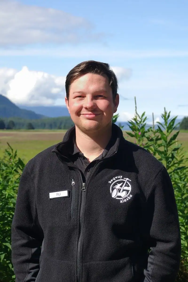 Get to Know Us 13 A smiling man wearing a black Segway Alaska fleece jacket outdoors with distant mountains.