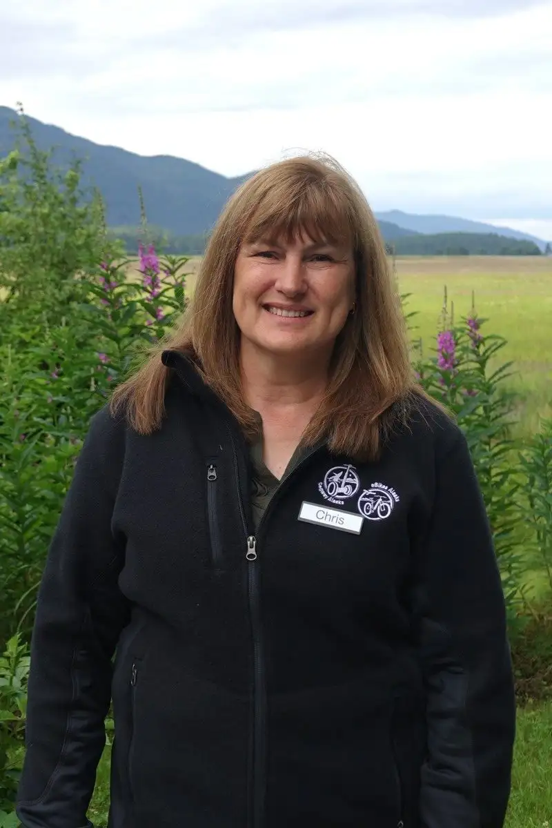 Get to Know Us 1 A smiling woman wearing a black fleece jacket outdoors with purple wildflowers and mountains behind.