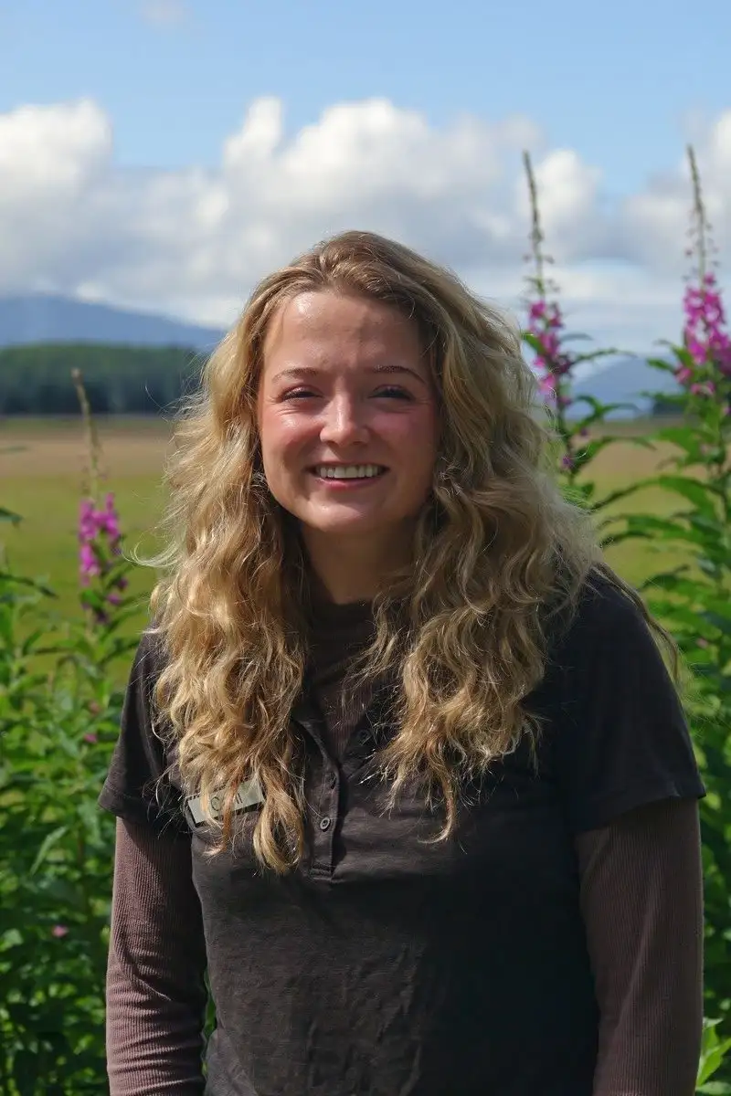Get to Know Us 4 A smiling woman with long, wavy blonde hair standing in a sunny field with wildflowers.
